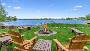 A lakeside deck with wooden chairs and tables, a fire pit, and a view of the lake. - Large Updated Lakefront House on Lake George. Kayaks, games, fire pit, sledding. (West Branch)
