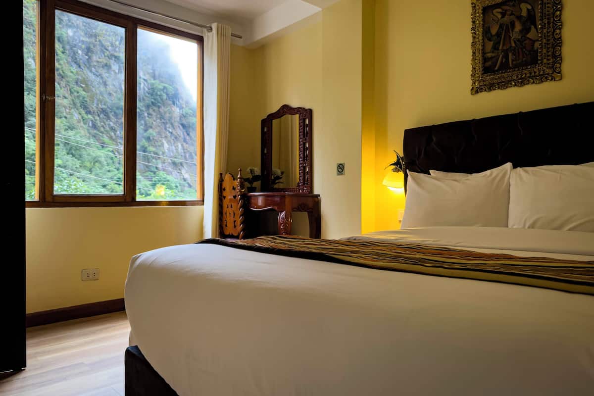 A hotel room with a large bed, a wooden nightstand, a mirror, and a view of a mountainous landscape through the window.
