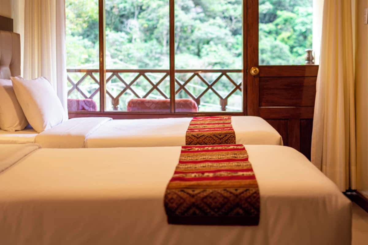 A hotel room with two beds, a wooden bench, and a view of greenery through large windows.