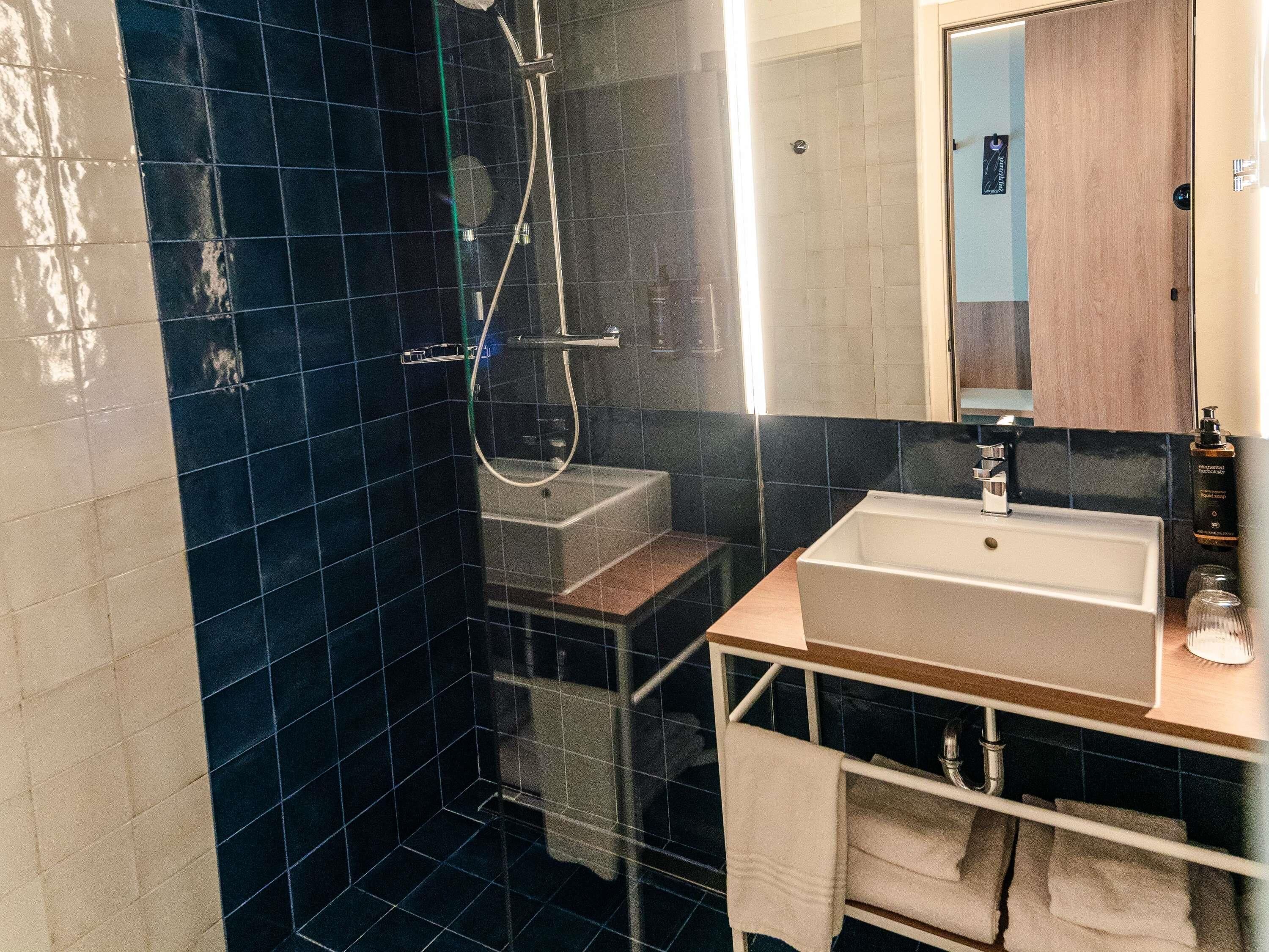 A modern bathroom with a dark blue tiled shower, a white sink, and a wooden vanity.