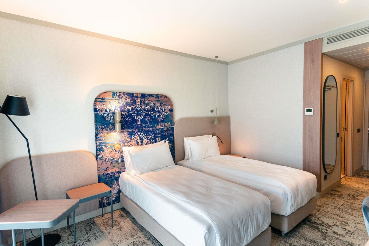 A hotel room with two beds, a desk, a chair, a large mirror, and a patterned headboard.