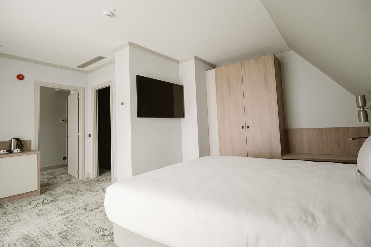 A modern bedroom with a large bed, a TV mounted on the wall, a wardrobe, and a desk with a kettle.