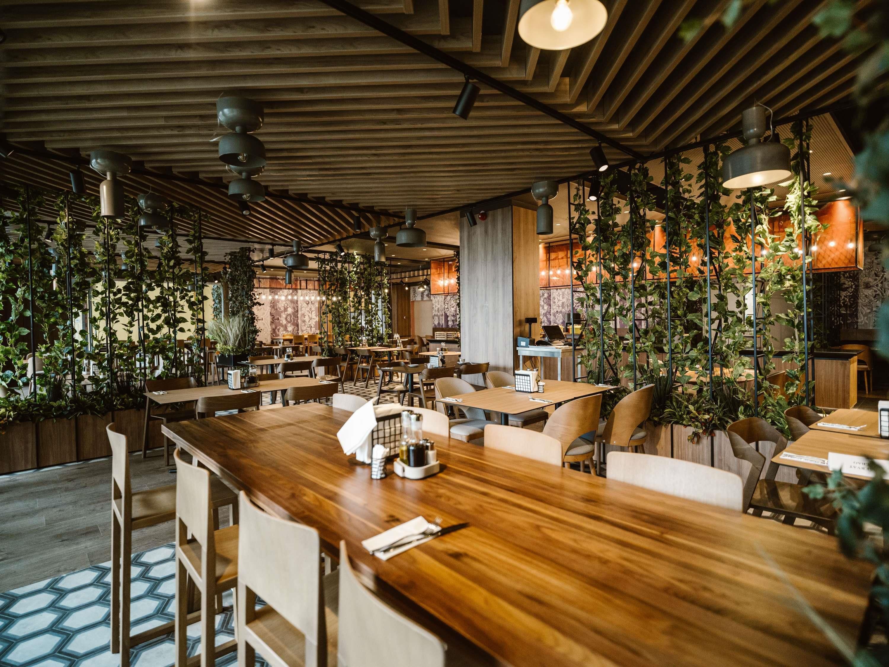 A modern restaurant interior with wooden tables and chairs, hanging plants, and pendant lighting.