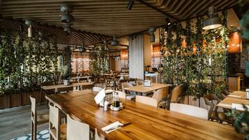 A modern restaurant interior with wooden tables and chairs, hanging plants, and pendant lighting.