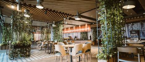 Un interior de restaurante moderno con suelo de madera, plantas verdes y una combinación de mesas redondas y cuadradas.