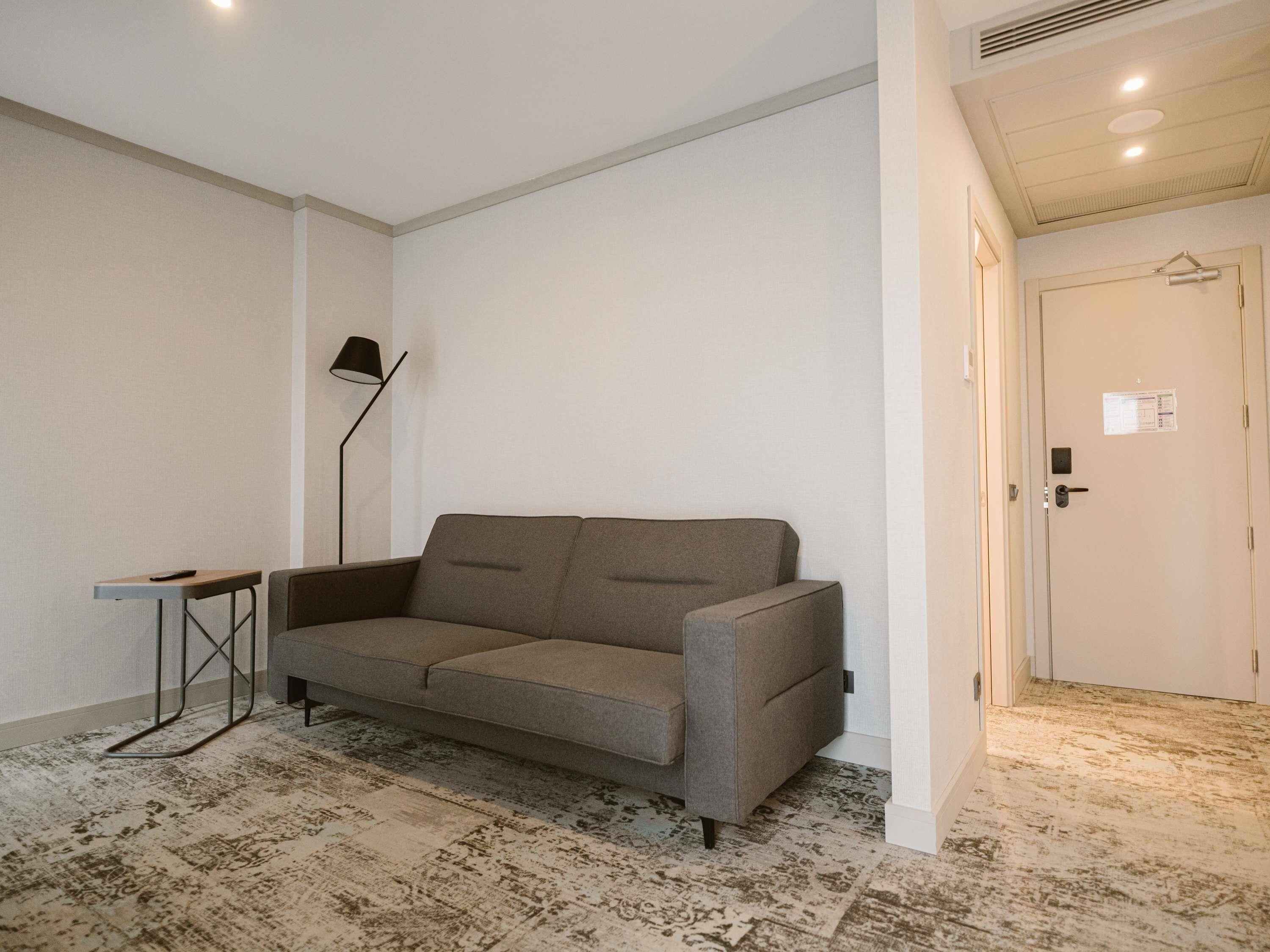A neutral-colored room with a grey sofa, a small side table, a floor lamp, and a door with a hanging coat rack.