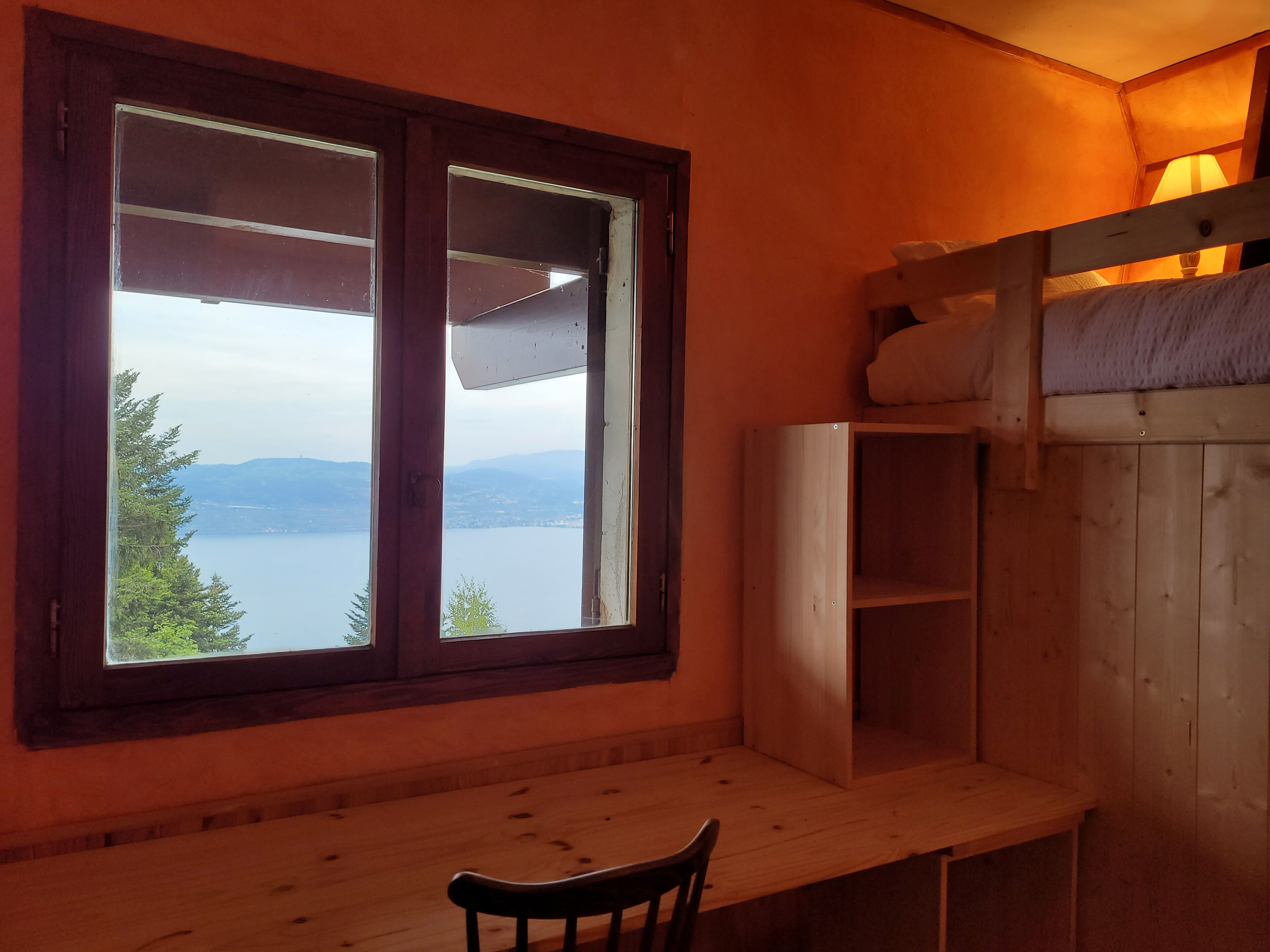 A room with a wooden desk, a chair, a bunk bed, and a window offering a view of a lake and mountains.