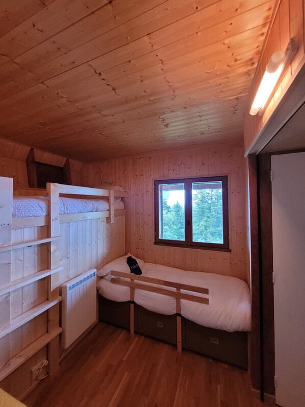 A compact wooden cabin room with bunk beds, a window with a view of trees, and a built-in heater.