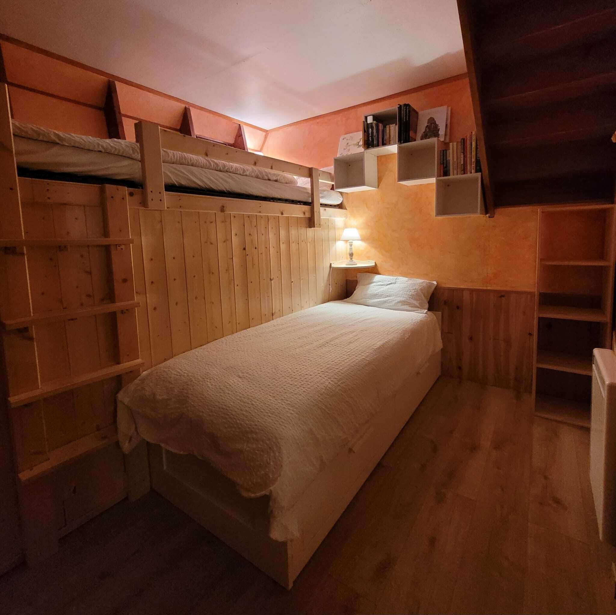 A cozy, compact room with a bunk bed, a bedside table with a lamp, and built-in shelves with books and decorative items.