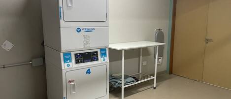 Een stapel van drie witte wasmachines met bedieningspanelen, een metalen plankenwand en een deur in een kamer met een effen muur en vloer.