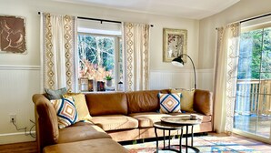 A living room with a brown leather sofa, a matching ottoman, a patterned rug, and a large window with curtains. - Jewel Brook Cottage- 4BR house on a tranquil flowing brook!  (Ludlow)