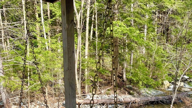 A wooden bench with a backrest and armrests, positioned on a gravel surface near a stream in a wooded area. - Jewel Brook Cottage- 4BR house on a tranquil flowing brook!  (Ludlow)