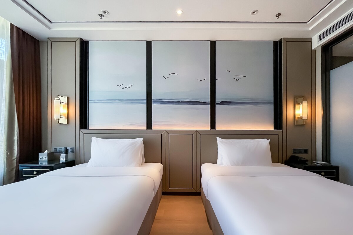 A hotel room with two beds, a large window with a view of the sea, and a nightstand with a lamp.
