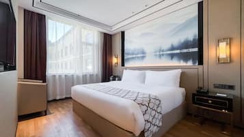 A hotel room with a large bed, a nightstand, a desk, a window with curtains, and a wall-mounted painting.