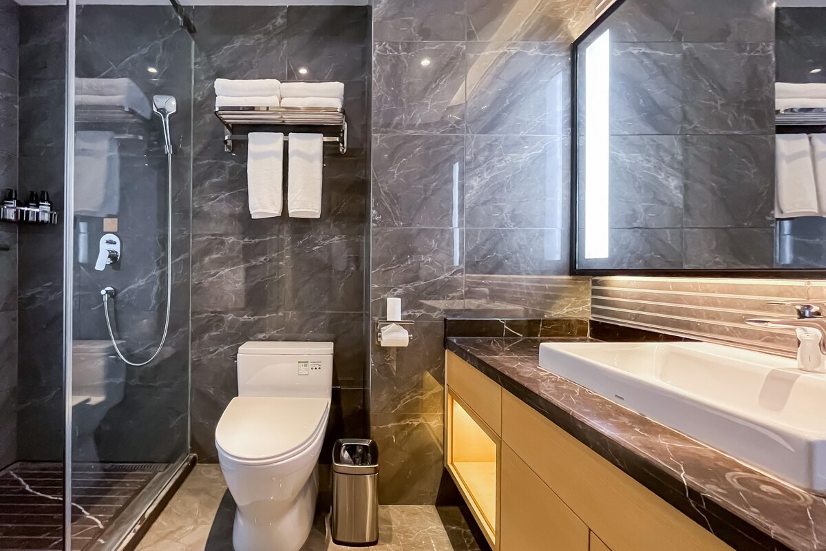 A modern bathroom with a glass-enclosed shower, a toilet, a trash bin, and a large mirror above a sink.