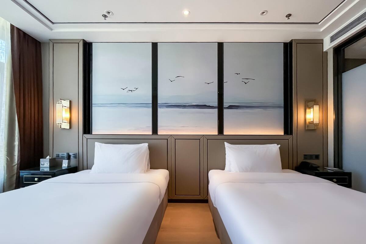 A hotel room with two beds, a large window with a view of the sea and birds, and a wall-mounted artwork.