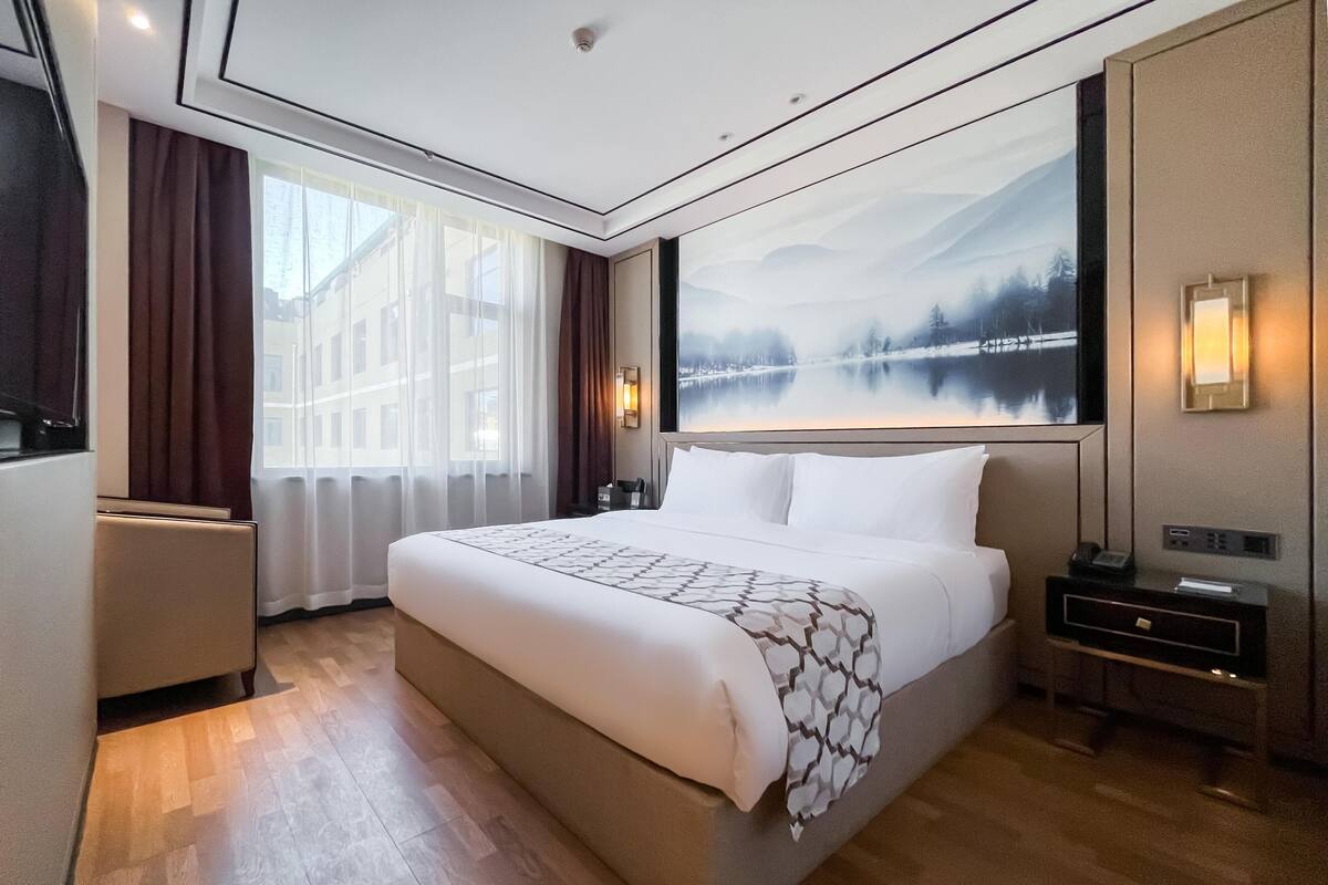 A hotel room with a large bed, a nightstand, a wall-mounted TV, and a painting of a lake.