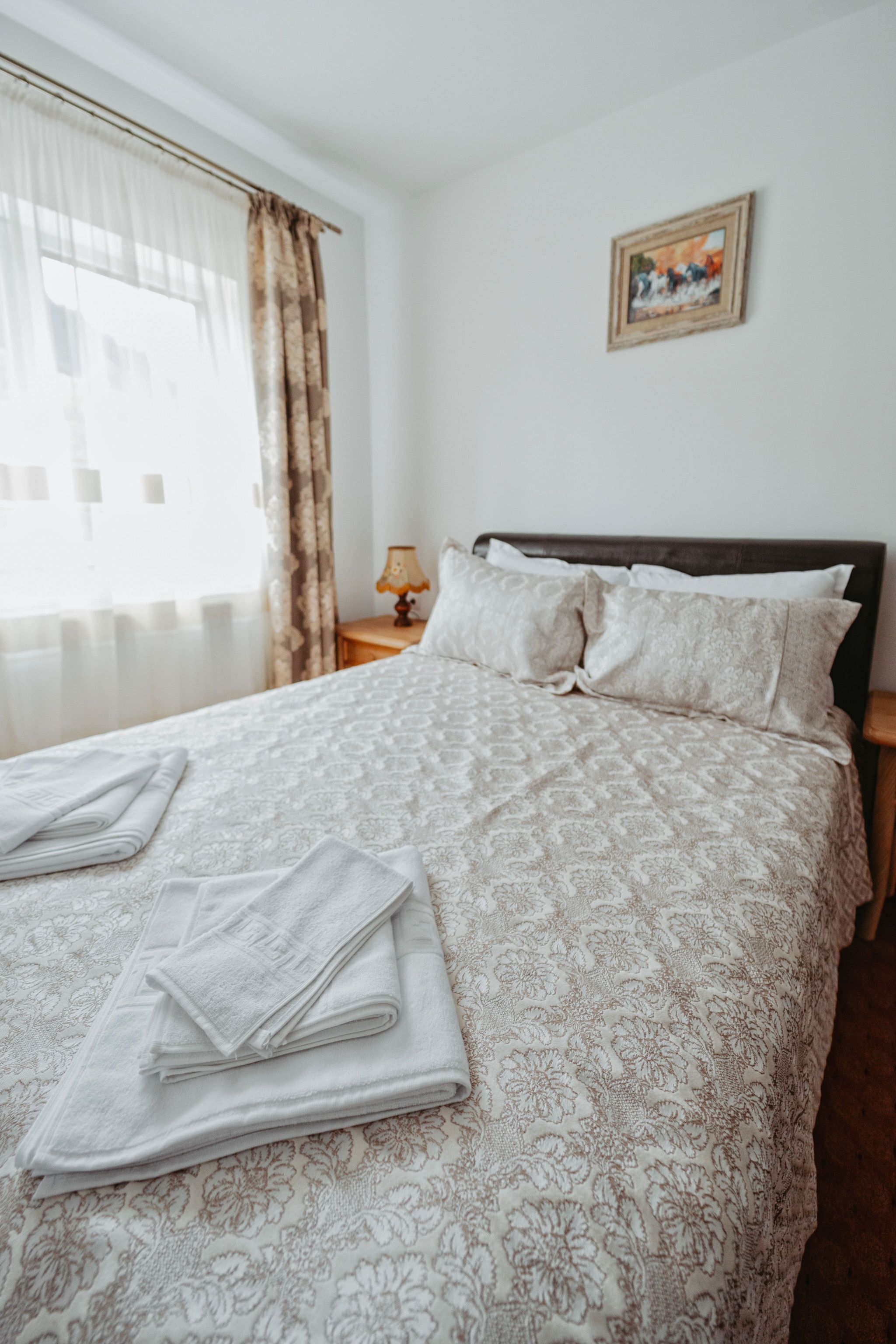 A neatly made bed with a patterned bedspread and pillows, a folded towel on the bed, a bedside table with a lamp, and a framed picture on the wall.