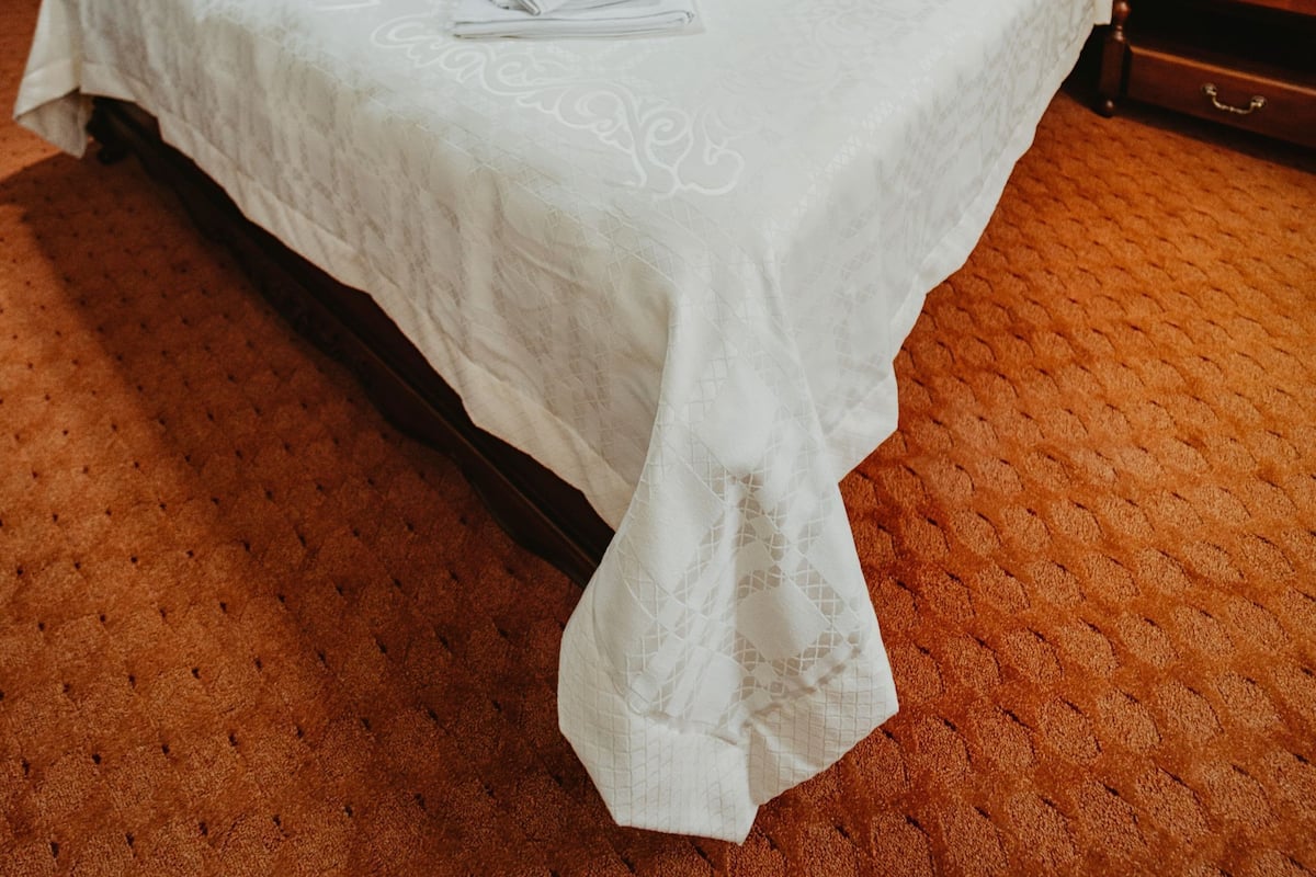 Un lit bien fait, recouvert d’une couvre-lit blanche, avec une tête de lit en bois et une table de chevet surmontée d’une lampe.