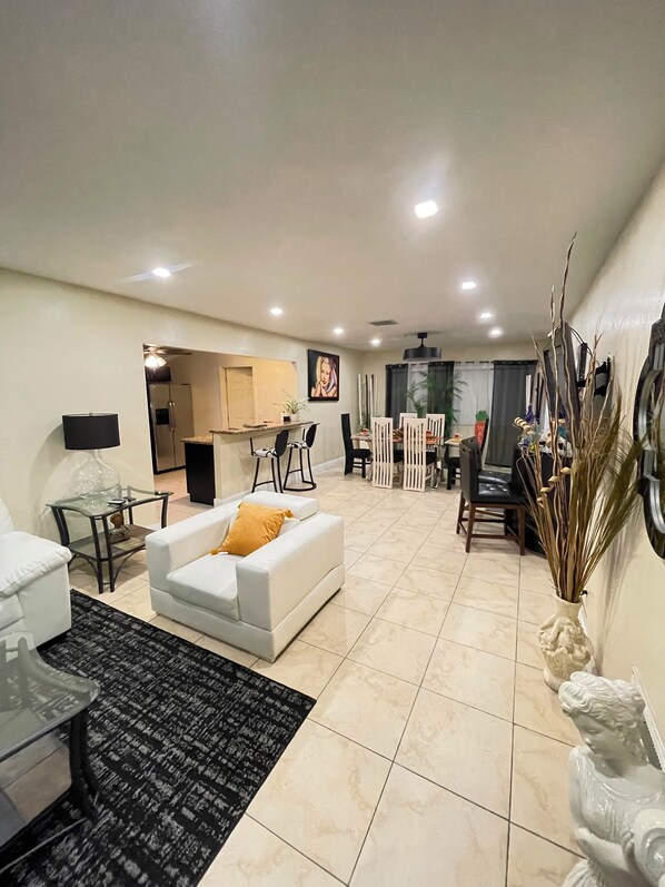 A modern living room with a white sofa, a glass coffee table, and a black rug. There is a dining area with a white table and chairs, and a kitchen in the background.
