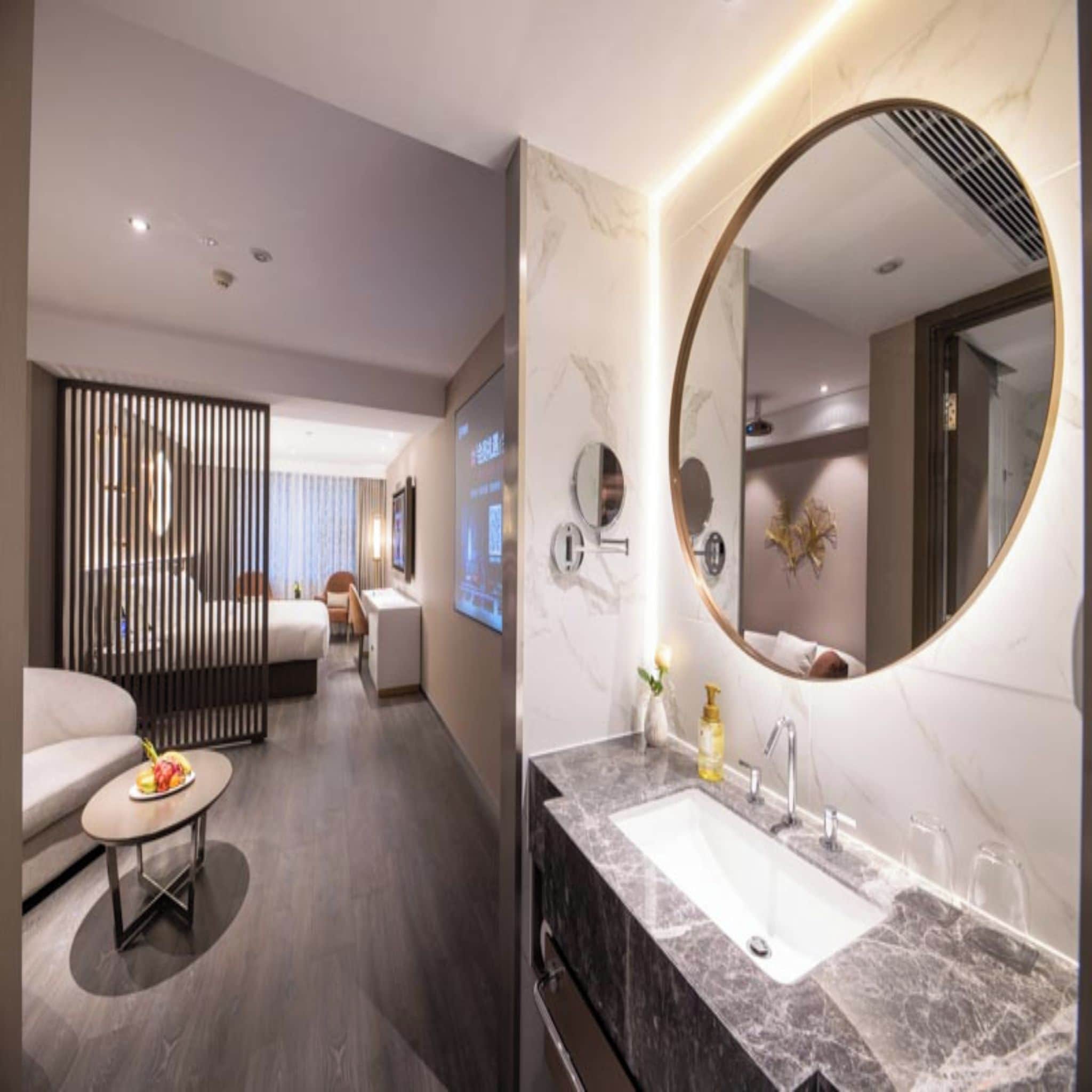 A modern hotel room with a large round mirror, a marble bathroom sink, a sofa, and a dining table with fruit.