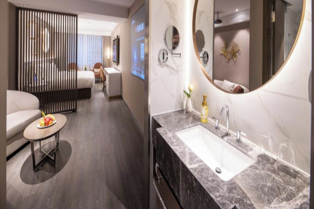 A modern hotel room with a large round mirror, a marble bathroom sink, a sofa, and a dining table with fruit.