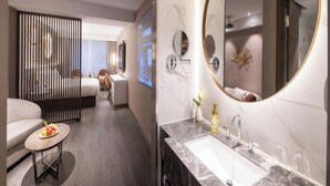 Quarto de hotel moderno com um espelho redondo grande, pia de banheiro de mármore, um sofá e uma mesa de jantar com frutas.