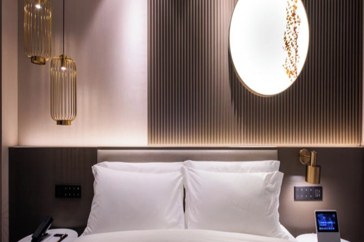 A modern hotel room with a large bed, two bedside tables, a wall-mounted circular light, and a wall panel with vertical lines.