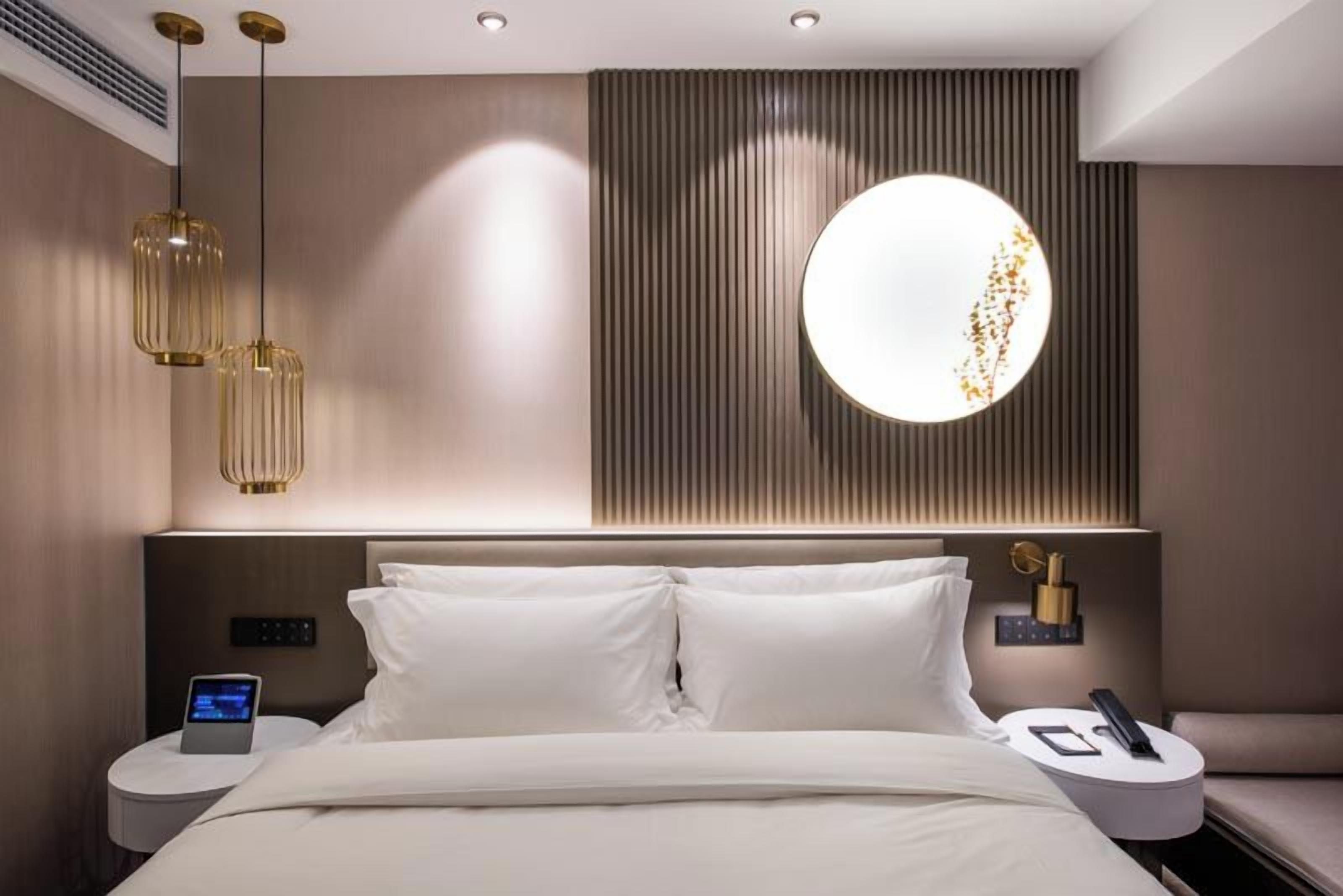 Una camera d'albergo moderna con un letto grande, due comodini, una luce circolare a parete e un pannello di controllo sul lato sinistro.