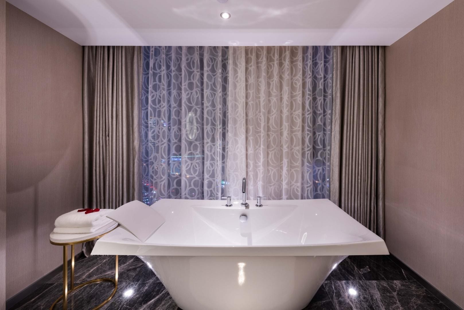 A modern bathroom with a freestanding bathtub, a vanity with a towel, and a view of the city through patterned curtains.