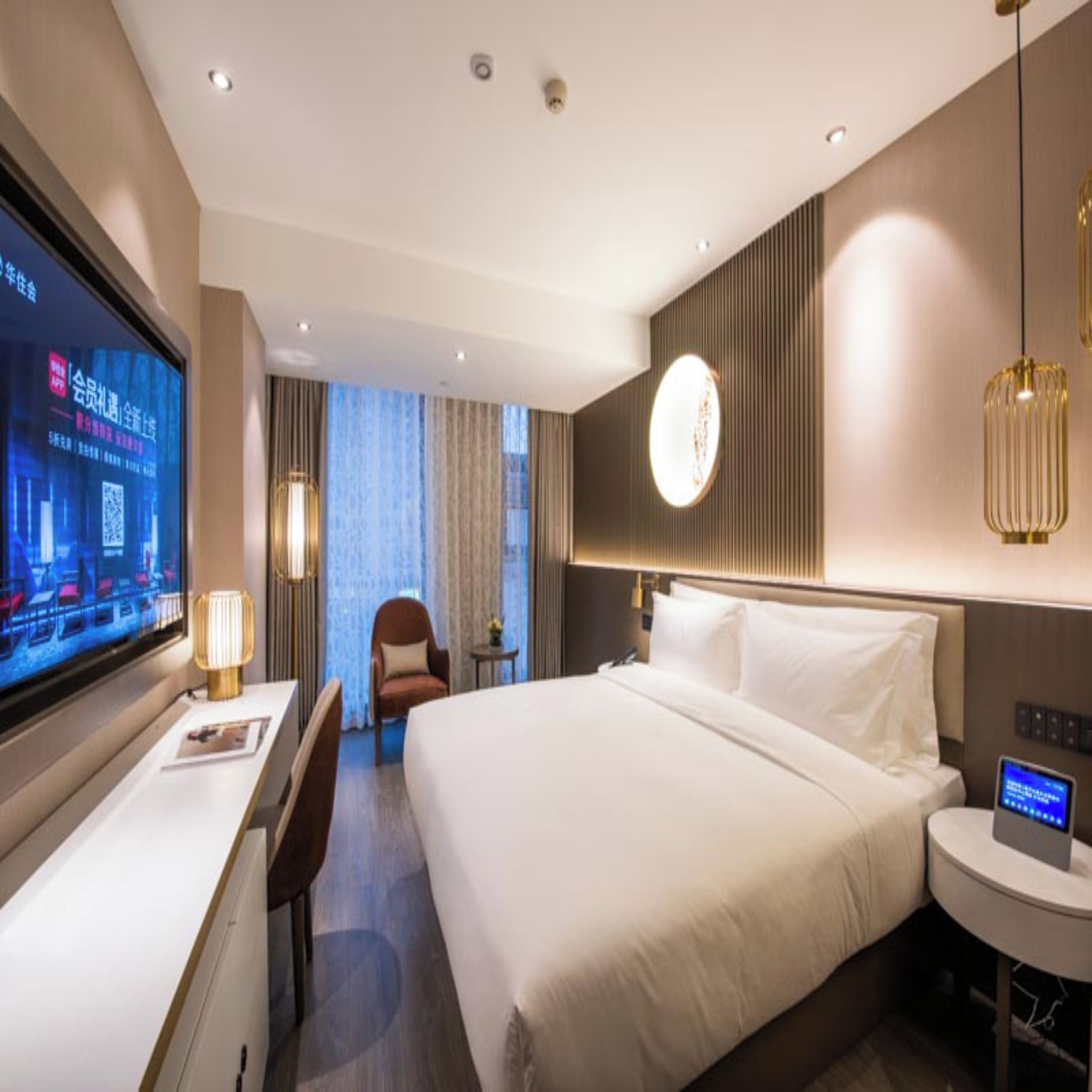 A hotel room with a large bed, a desk with a computer, a chair, and a TV displaying a QR code.