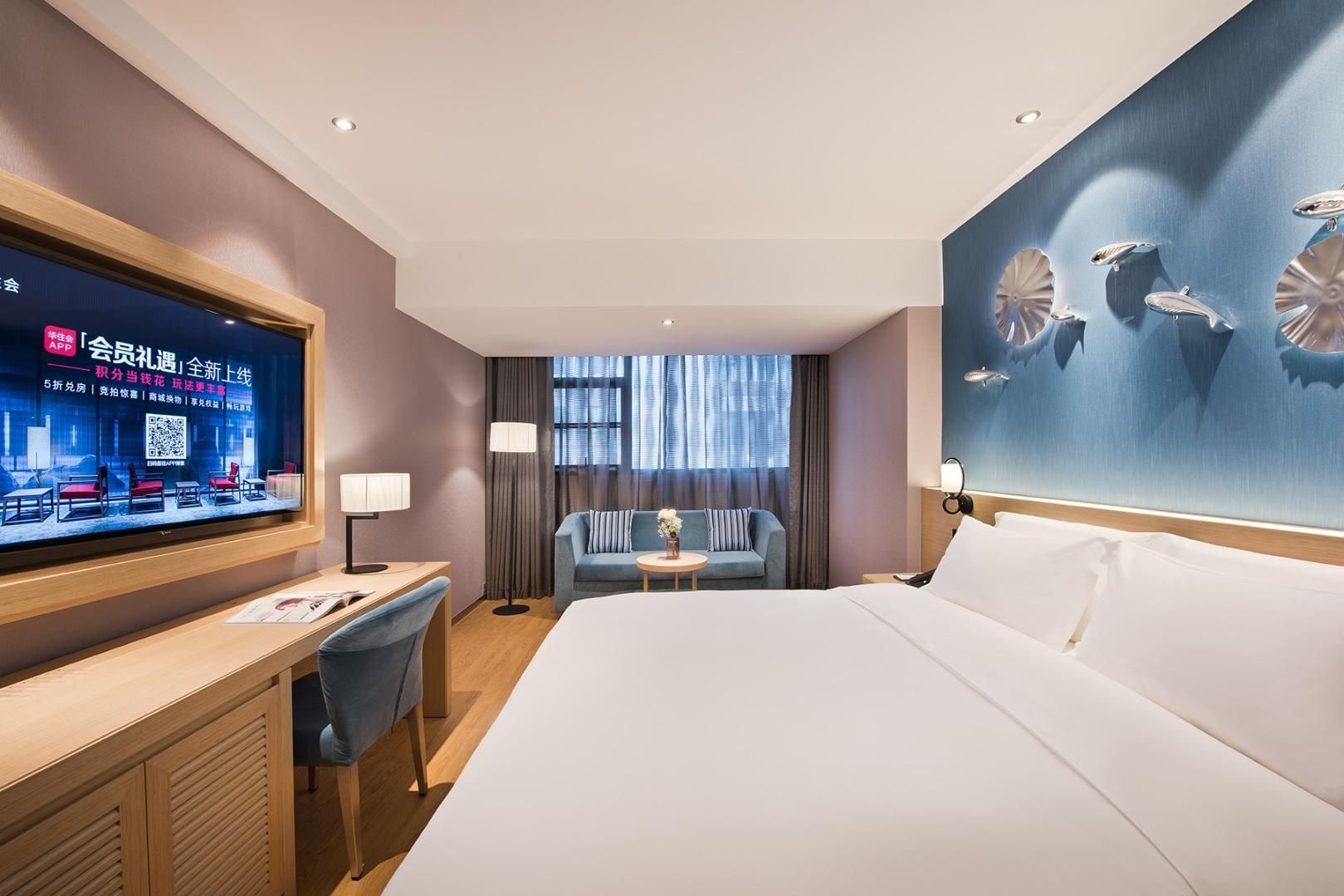 A hotel room with a large bed, a desk with a chair, a small table with a lamp, a sofa, and a TV mounted on the wall.