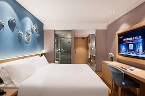A hotel room with a large bed, a desk with a chair, a television, and a glass-enclosed shower area.