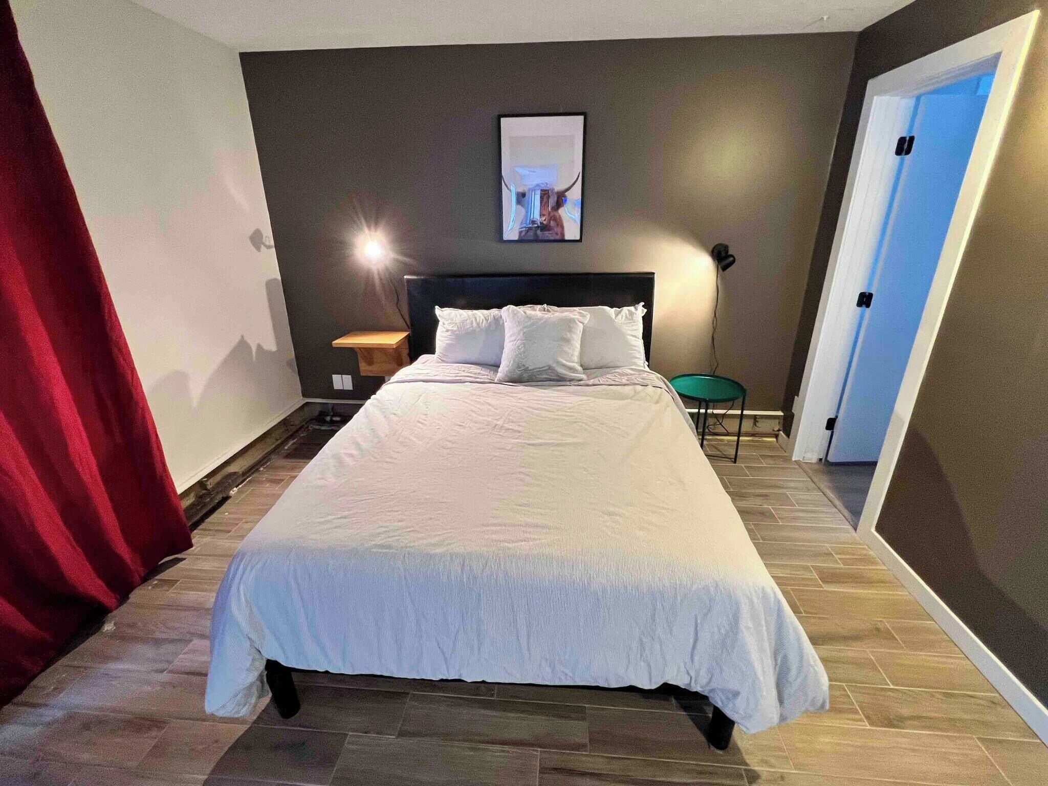 Ein Schlafzimmer mit Bett, Nachttisch, Stuhl, einer Tür und einem Wandbild.
