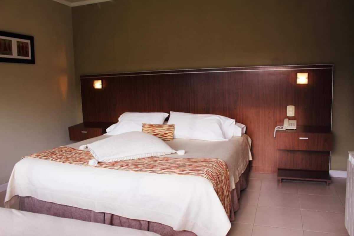 A hotel room with a large bed, a wooden headboard, bedside tables with lamps, and a small wooden shelf with a telephone.