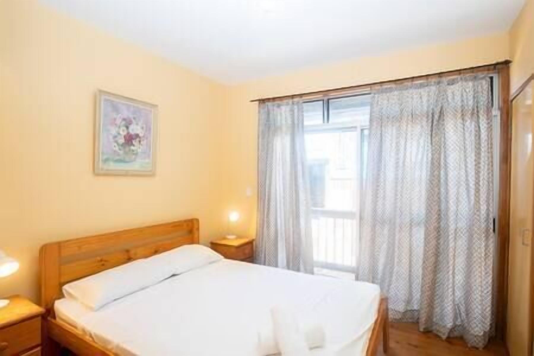 Een slaapkamer met een houten bed, een nachtkastje, een lamp, een schilderij aan de muur, en een raam met gordijnen.