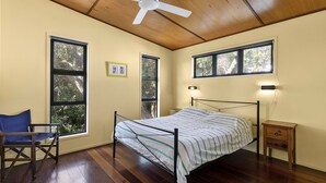 Une chambre à coucher comprenant un lit, une chaise, deux fenêtres, un ventilateur de plafond et une table de chevet.
