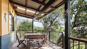 Un espace extérieur abrité, doté d’une table et de chaises en bois, d’une rambarde et offrant une vue sur les arbres et les collines.