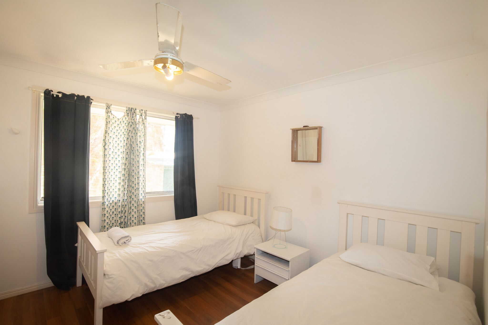 A small, clean room with two single beds, a ceiling fan, a window with curtains, and a bedside table with a lamp.
