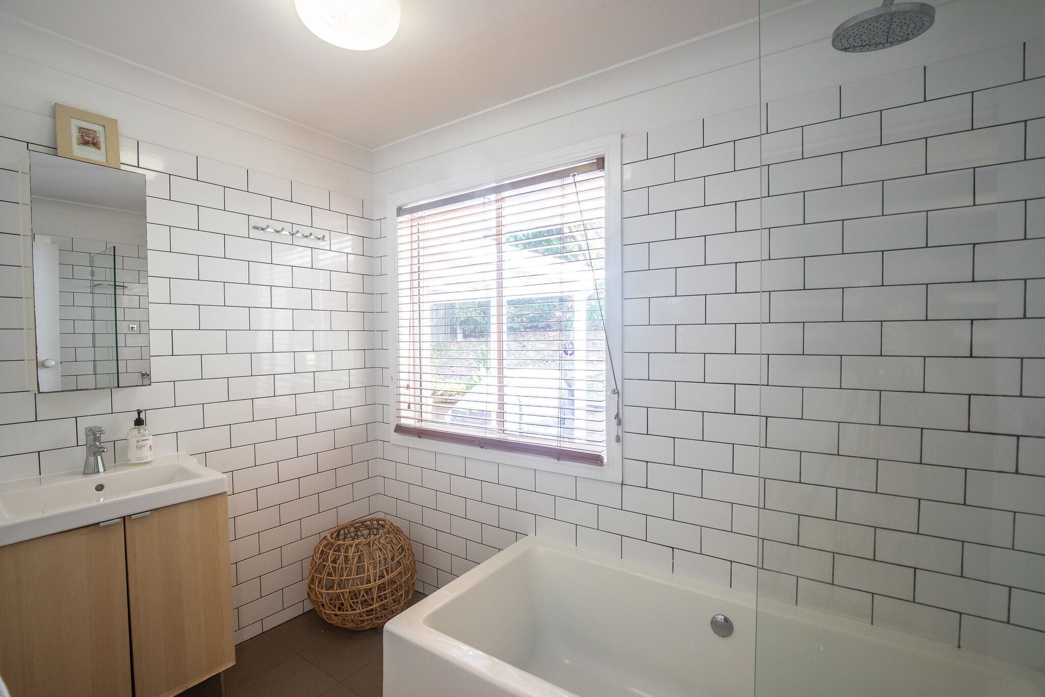 A bathroom with white tiled walls, a white bathtub, a sink with a mirror, and a window with blinds.