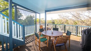 Um terraço coberto ao ar livre com mesa e cadeiras de madeira, churrasqueira e vista para árvores e colinas.