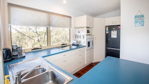 Uma cozinha moderna com armários brancos, bancada azul, pia de aço inoxidável, geladeira preta e janela com vista para árvores.