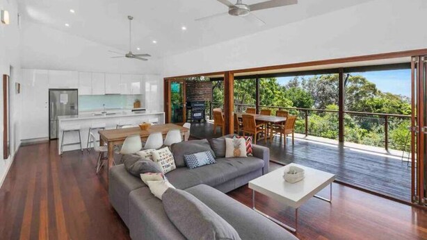 A modern living room with a grey sofa, dining table, and chairs, a kitchen area, and a balcony with a view of greenery. - This architect-designed Straddie beauty is the perfect laid-back beach house (Point Lookout)