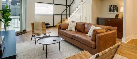 A spacious living room with a brown leather sofa, a coffee table, and a chair. A staircase leads to a second floor, and there is a large window with blinds.