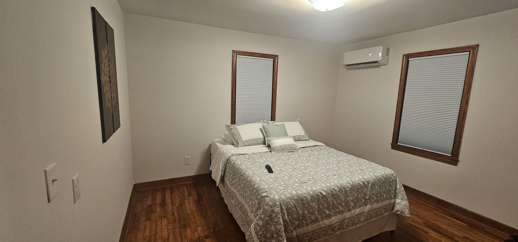 A bedroom with a bed, two windows with blinds, an air conditioning unit, and a wooden floor.