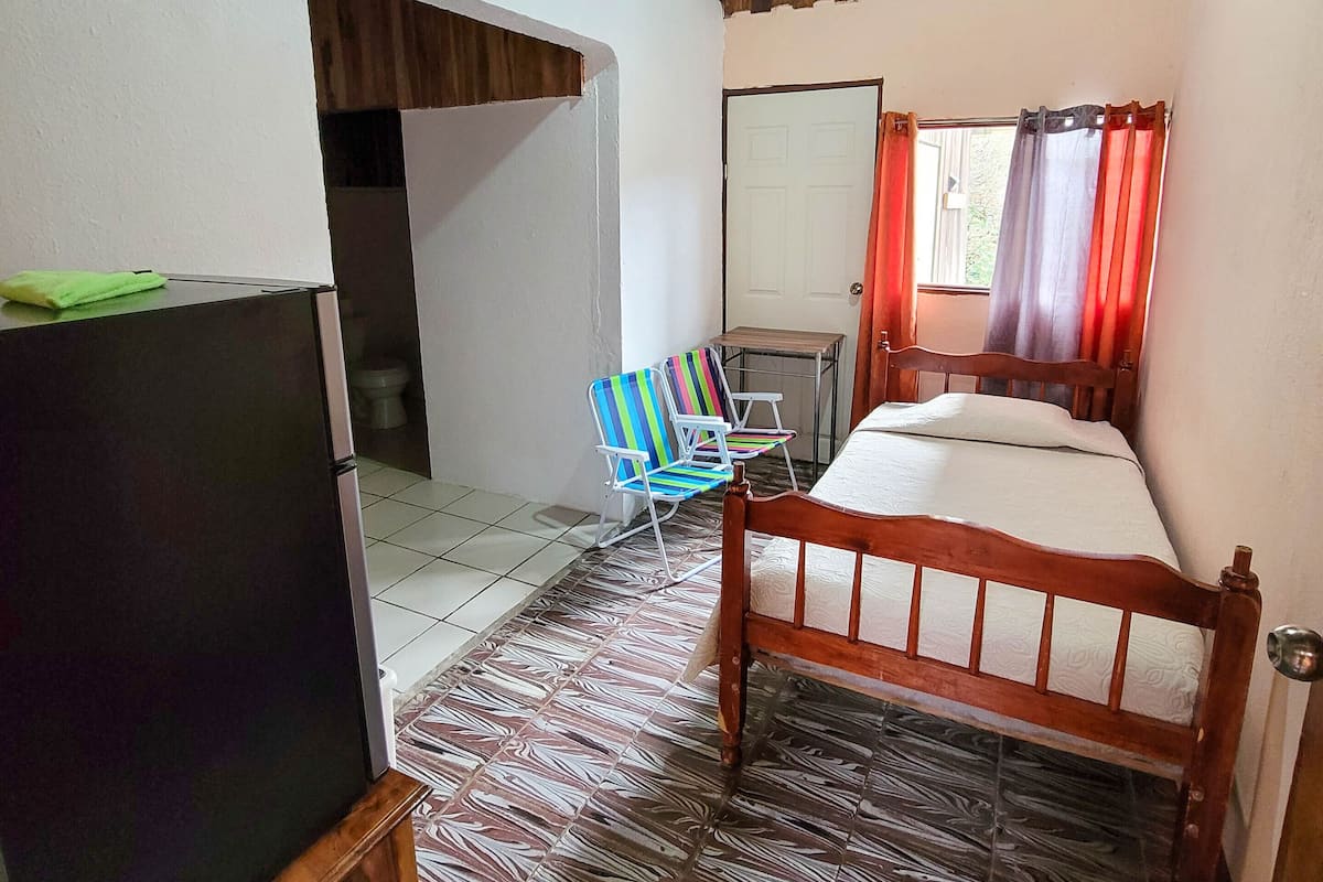 A room with a wooden ceiling, a bed, a TV, a chair, and a window with curtains.