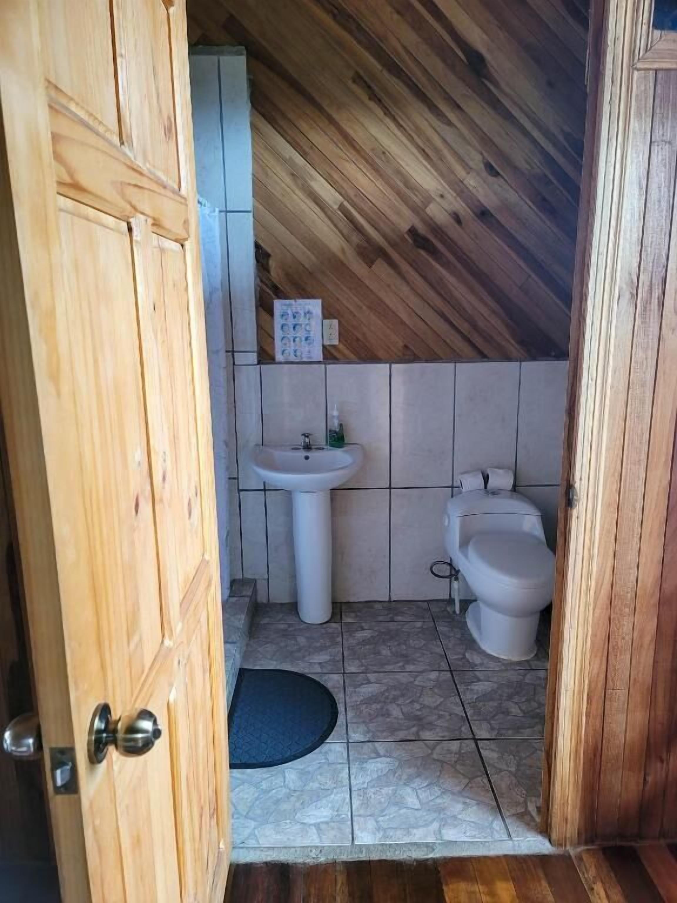 Um banheiro pequeno com teto de madeira, pia branca, vaso sanitário e piso de azulejos.