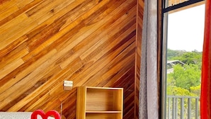 A room with a wooden sloped ceiling, a bed with a heart-shaped decoration, a wooden shelf, and a window with a view of greenery. - Cabinas Selva Verde (Monteverde)