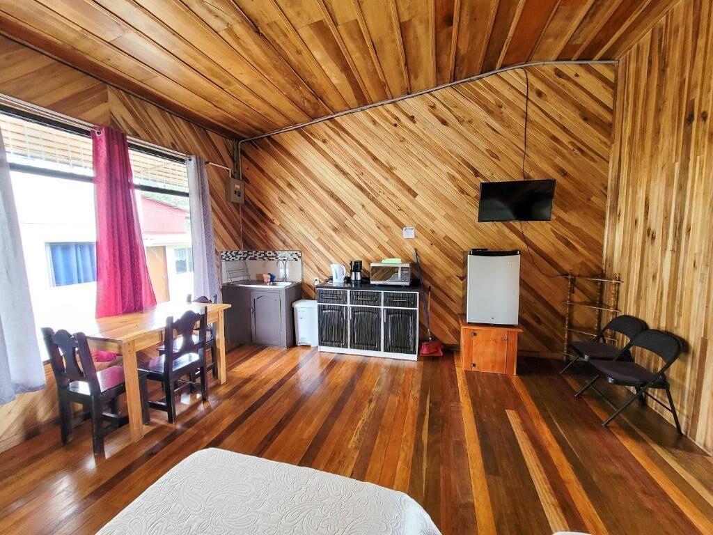 A room with wooden walls and a sloped ceiling, a dining table with chairs, a bed, a TV mounted on the wall, and a small kitchenette area.