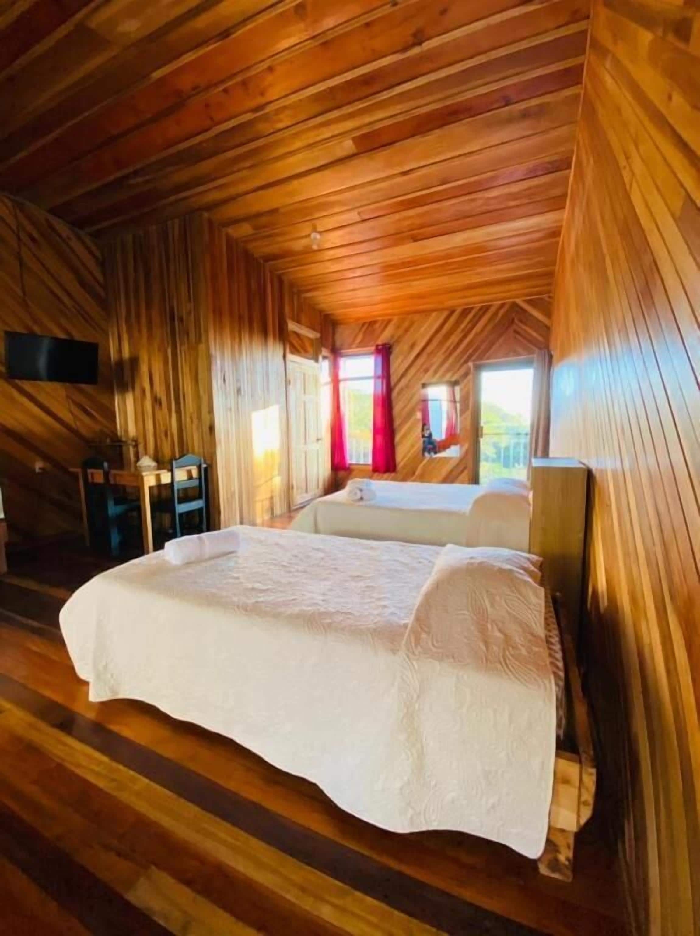Un dormitorio de cabaña de madera con una cama, un televisor montado en la pared y una ventana con cortinas.